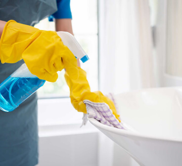 Close-up of hands in yellow gloves cleaning a white bathroom sink with a purple cloth and a spray bottle, representing housekeeping and cleaning tasks.