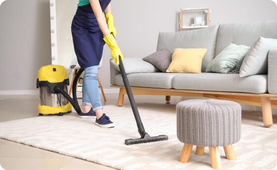 Person vacuuming a carpet in a living room with a grey sofa and a small ottoman, using a yellow and black canister vacuum cleaner.