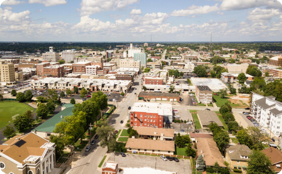 Aerial view of a small town with mixed residential and commercial buildings, streets lined with trees, and scattered parking areas on a sunny day with a clear sky.