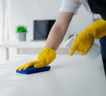 
A person in yellow gloves wiping a white surface with a blue cloth, with a spray bottle in the other hand, in a clean, white-themed room.